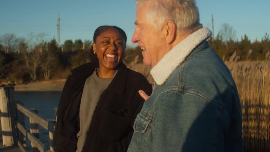 A young woman and an elderly man stand on a dock and talk and laugh together