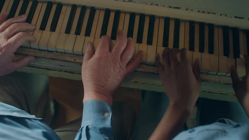 Elderly man with blue shirt and woman with bun and blue shirt play piano together