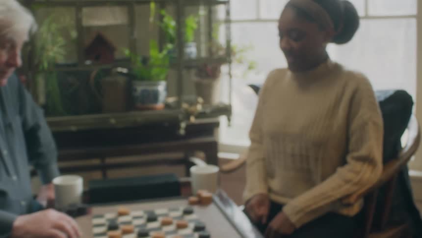 A man and woman are sitting in a home during the day playing checkers