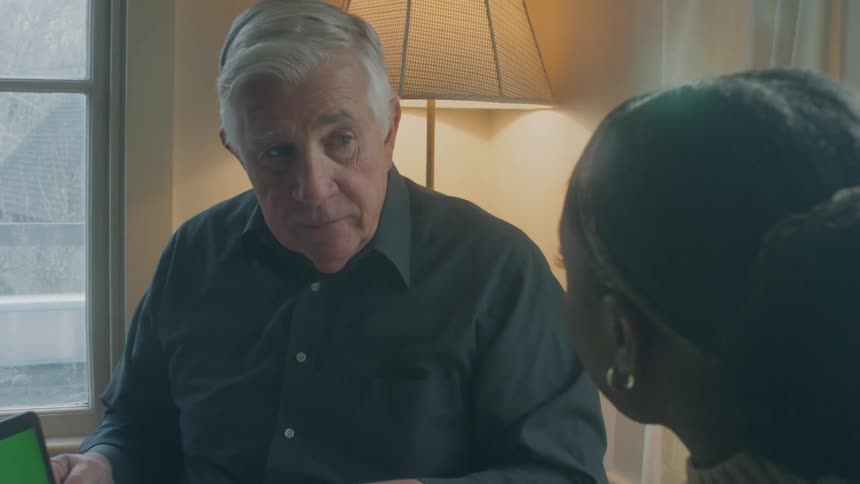 Old man sitting indoors next to a window holding a laptop and talking about it with a black woman