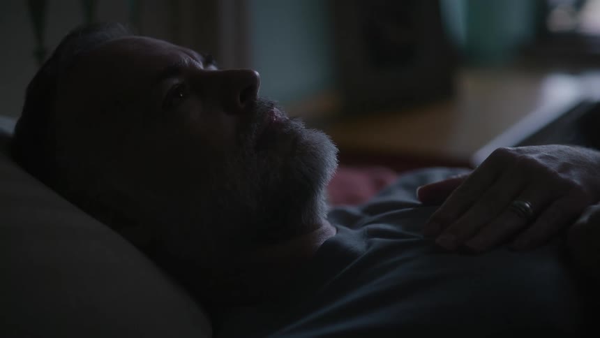 Man lies down onto bed hand to his chest then after staring at the ceiling sits back up