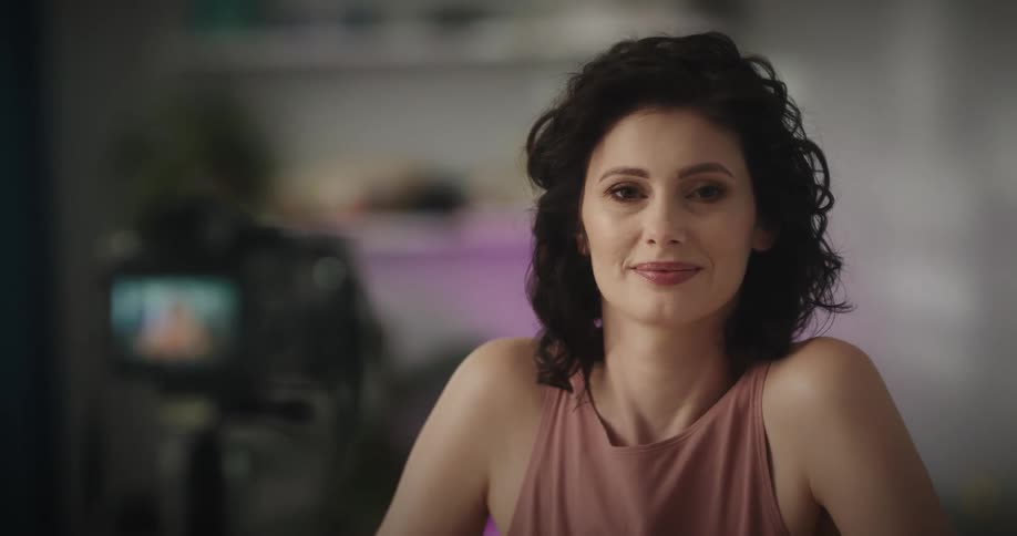 A woman puts down the makeup shes holding and smiles at the camera on a tripod
