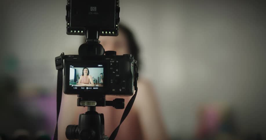 A woman walks into a room at home and turns on a camera mounted on a tripod and sits down to begin recording video in the daytime