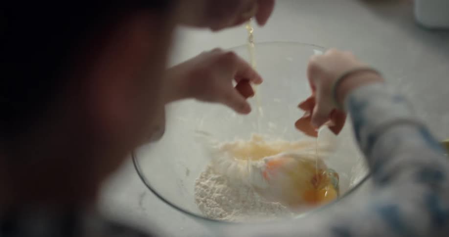 A woman and a girl are both cracking eggs into a bowl containing flour in the kitchen in the morning
