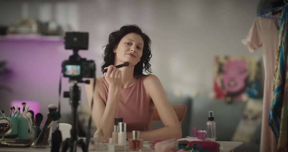 A woman is sitting indoors at a table in front of a camera recording herself demonstrating how to use a makeup brush