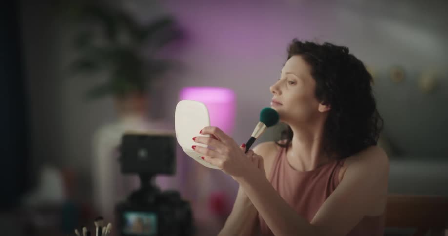 A woman is sitting in front of a video camera recording herself applying makeup with a brush in the evening