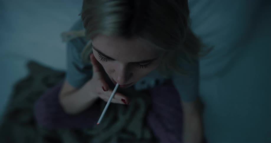 Blonde woman with gray and purple pants sits on bed and lights a cigarette at night in bedroom