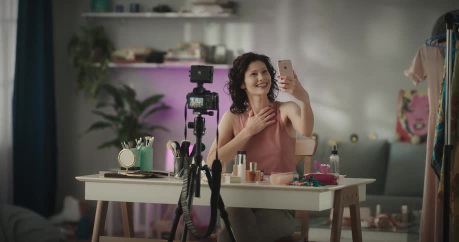 Woman sits at a table that is covered with makeup items and looks at her cellphone and smiles and laughs