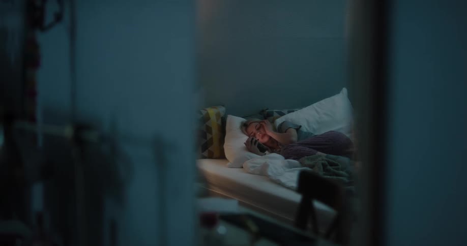 Blonde woman in gray t shirt and purple pants lies in bed and uses smartphone in bed in bedroom