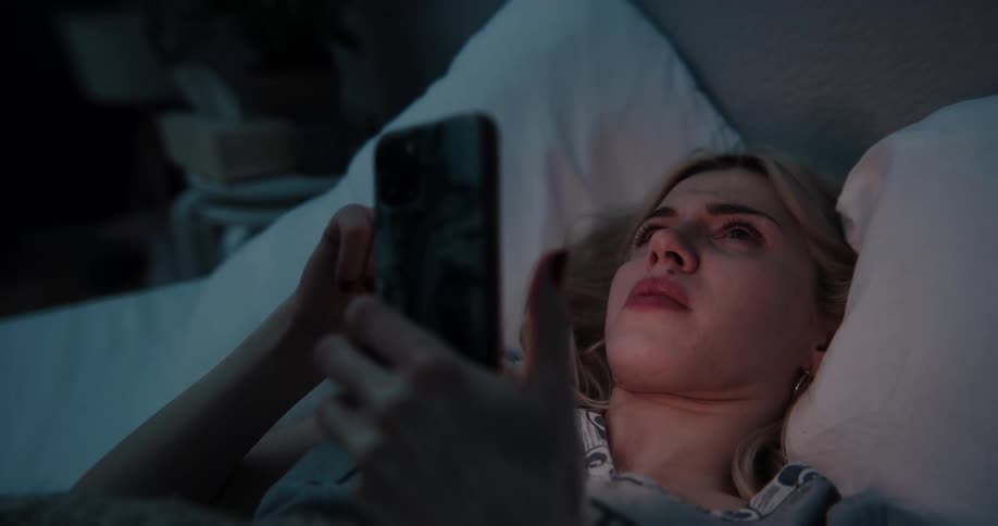 Sad young woman scrolling on her smartphone while laying in bed at night