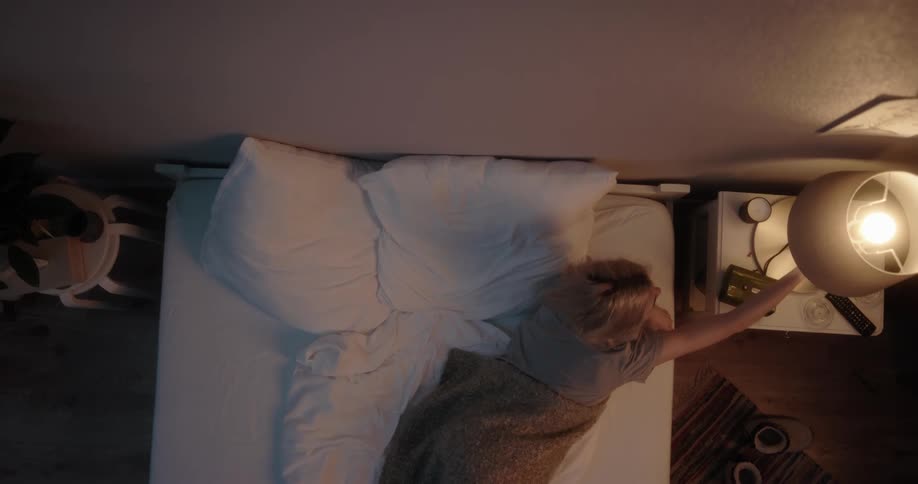 Young white woman in her bedroom lying in the bed then yawning turning off the table lamp and going to sleep