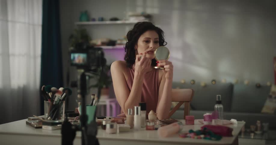 Woman sitting at a desk in her living room records a makeup tutorial while talking to the camera