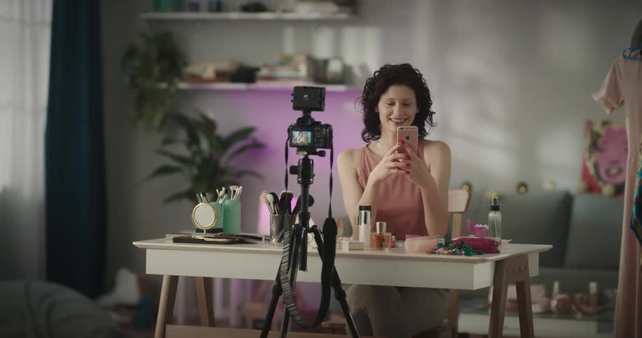 Woman views herself in her smartphone and talks as camera on tripod tapes her