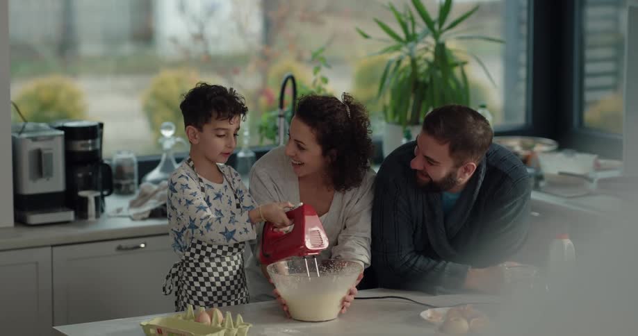 Mom enters the kitchen where dad and son are making breakfast and holds the bowl while son mixes