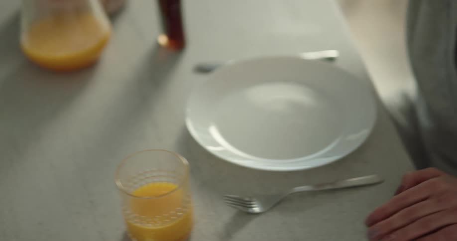 A woman covering her eyes sits down at a table as a man slides a plate of pancakes towards her