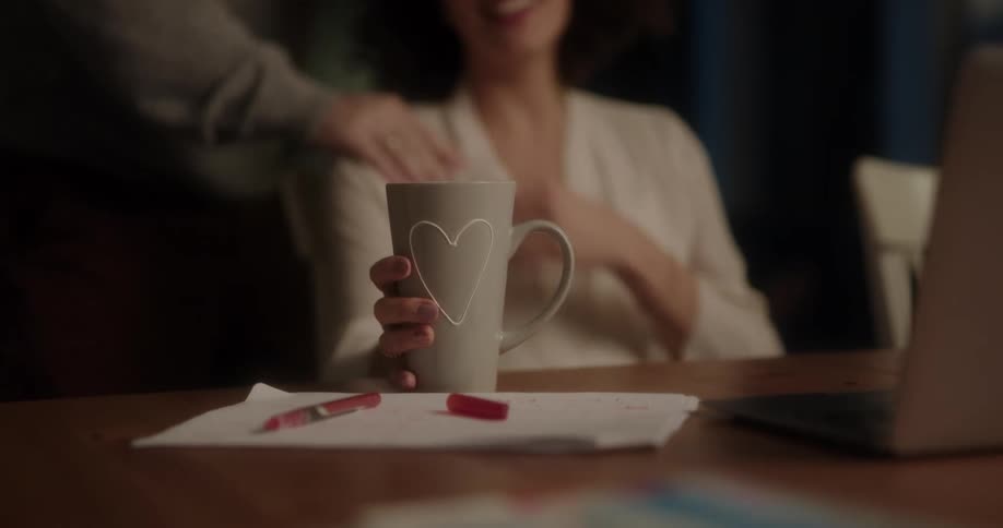 Man brings a woman a drink in a mug as she laughs and looks at a laptop