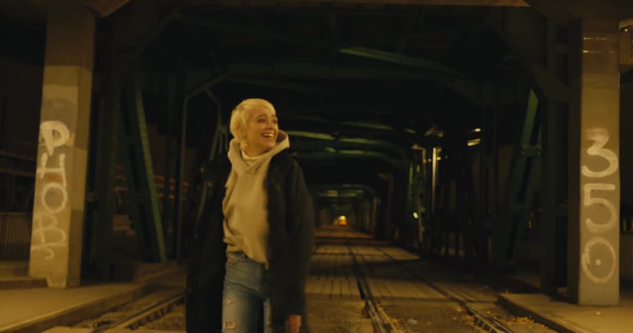 A teenage girl laughs and spins around and throws her arms in the air as she walks down train tracks
