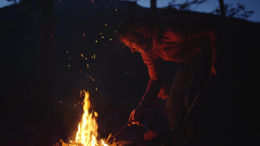 A man leaning over the flames stoking a campfire with a stick