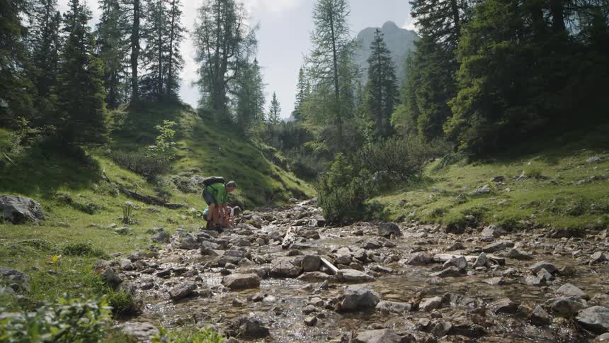 Family of four hiking on the mountains stops by a shallow river to drink water and wash