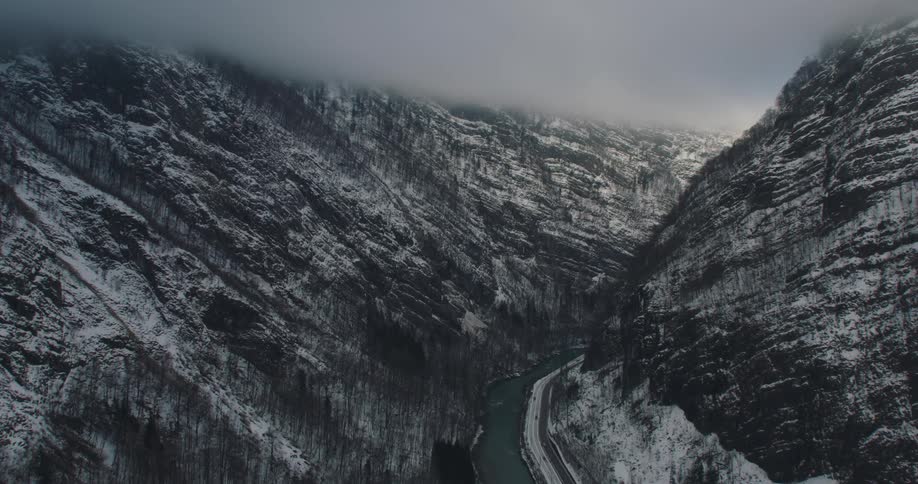 Aerial view of a road on the bottom of a canyon in the mountain next to a river under the clouds