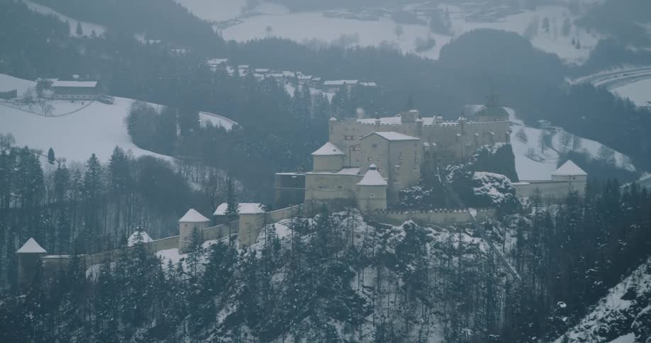 Aerial view of a castle on the mountains surrounded by pine tree forests covered in snow
