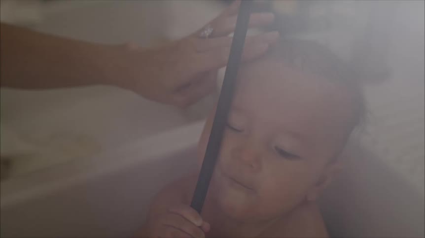 Baby tries to put a sprayer hose in his mouth while mom gives him a bath in the sink