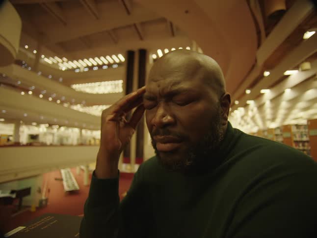A man sitting in a library rubbing his head and grimacing as he thinks