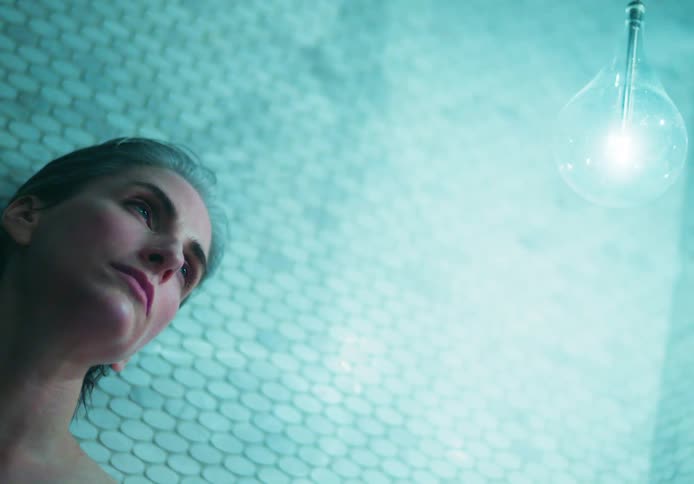 A woman leaning against a tile wall touches her damp hair and looks at a bright bulb