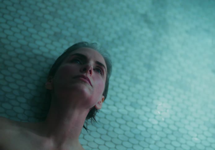 A woman leaning against a tile sauna wall wipes her face with her hands