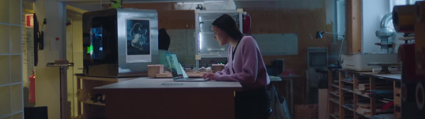 Woman working inside a workshop in front of a laptop near 3d printers and shelves filled with materials
