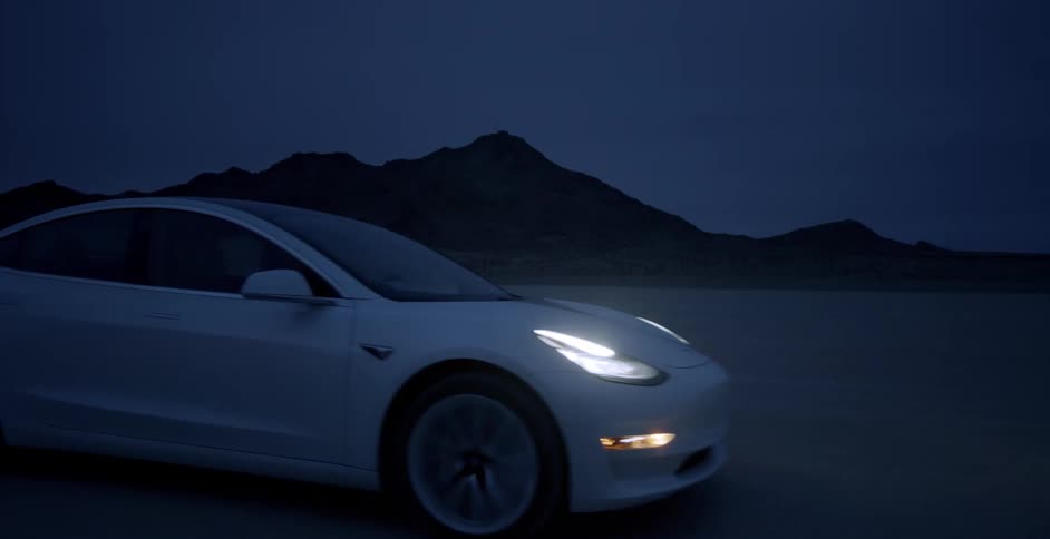 A car is traveling in a flat desert terrain at night with a mountain silhouette in the background