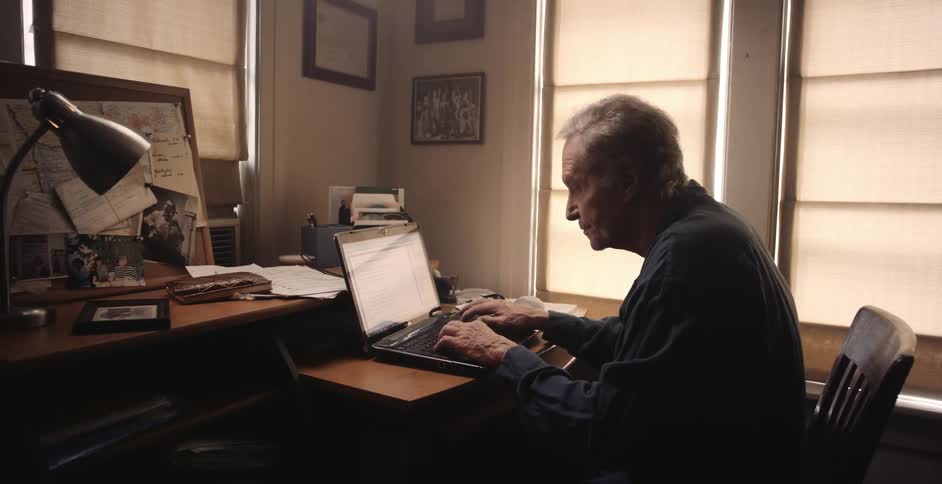 An elderly man is typing on a laptop indoors