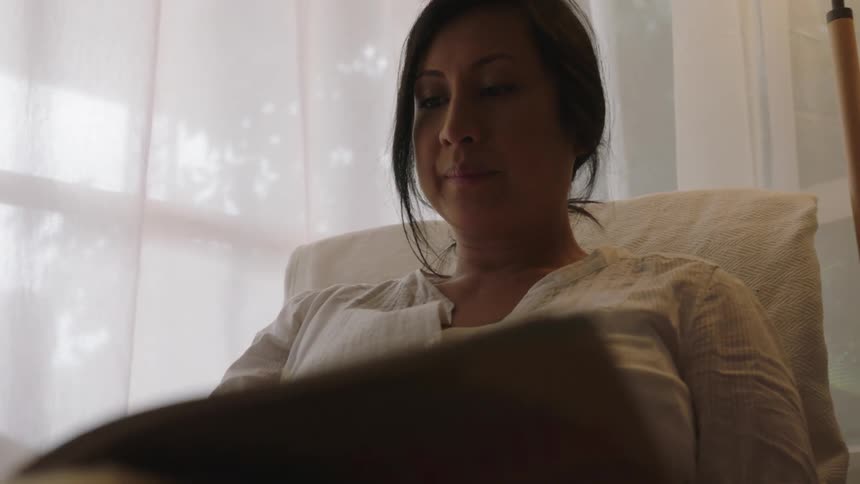 Woman with white dress sits in armchair and turns page next to windows
