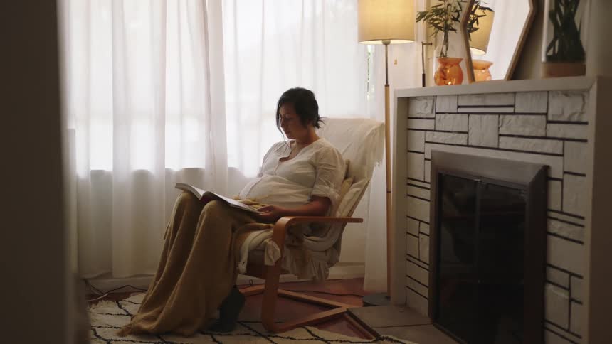 A pregnant woman is reading a book and sitting indoors