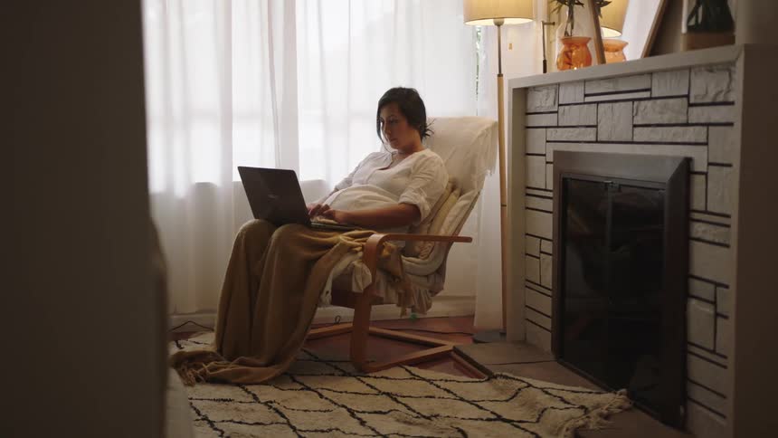 Pregnant woman with a laptop on her lap sitting on a chair at home next to a window