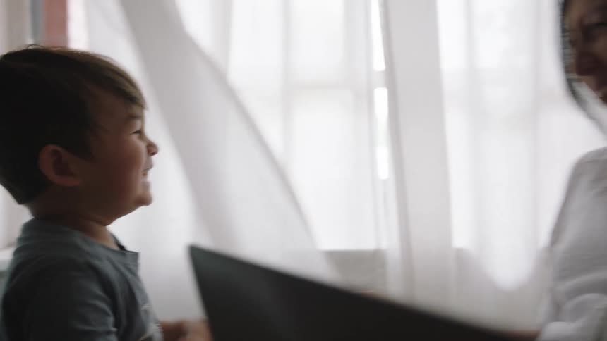 A boy hides behind sheer curtains and plays with his pregnant mom while she sits with a laptop