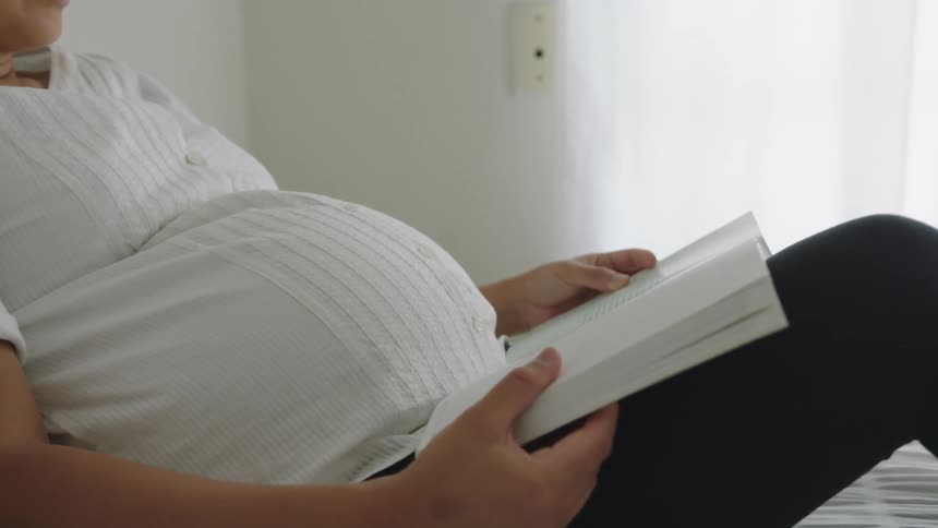 A pregnant woman touches her belly as she reads a book in bed