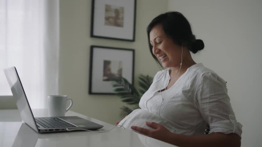 Pregnant woman with headphones having a facetime chat with laptop at dining table