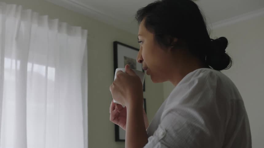 Pregnant woman with white shirt drinks from cup and looks out the window
