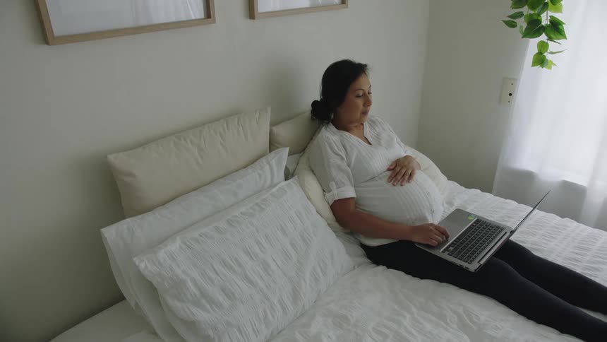 A pregnant woman leans back as she works on her laptop in bed