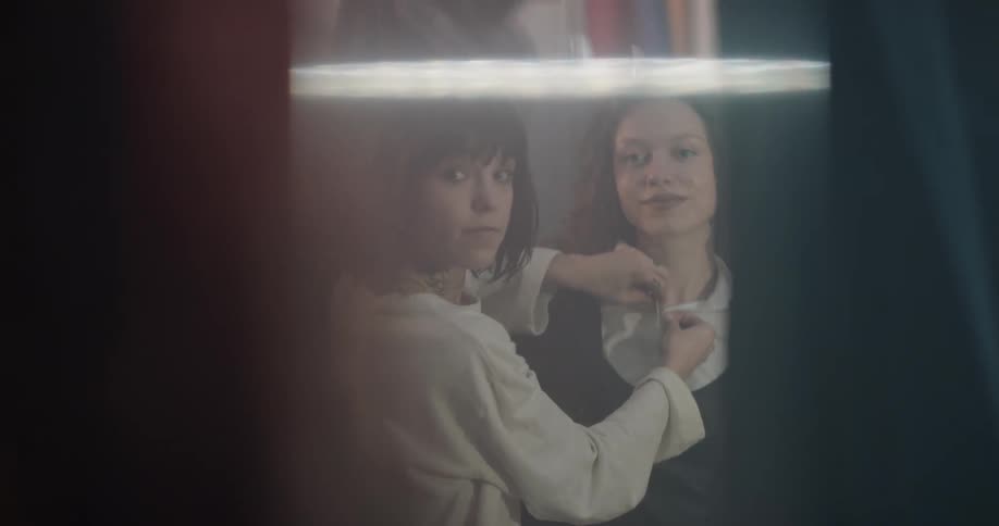 A stylist helps a teenage model get dressed as they look in a mirror