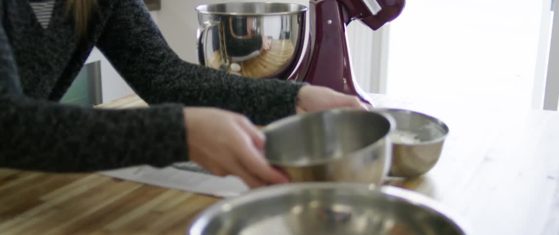 A woman lifts a bowl and looks at a recipe while in a kitchen