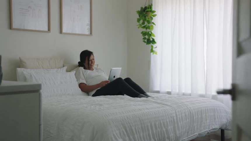 Pregnant woman reclines on bed using earphones with her tablet and laughs while stroking her belly