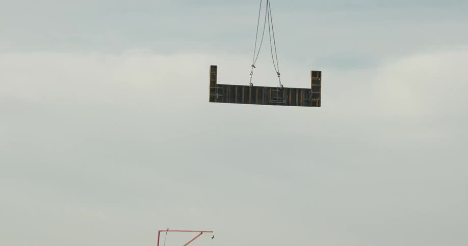 A construction crane is lowering a large piece of building material to the construction site and the crew below