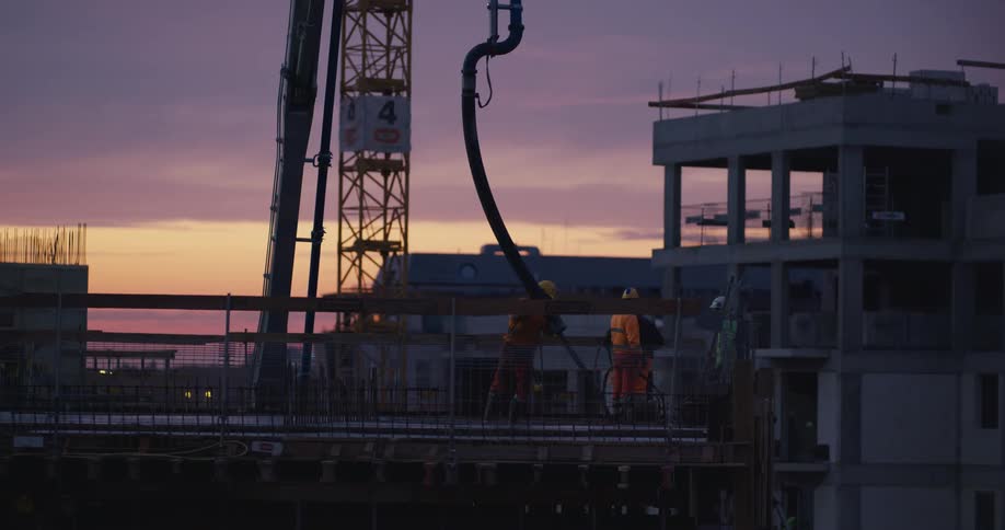 Men spread concrete along floor of building under construction as water in the background reflects the red sky