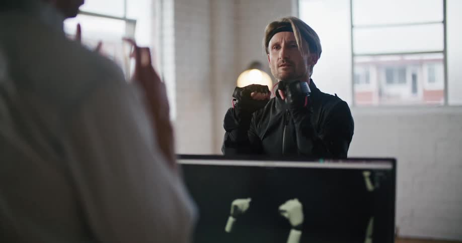 A man wearing virtual reality devices is speaking with a man sitting behind a computer and imitating his hand movements