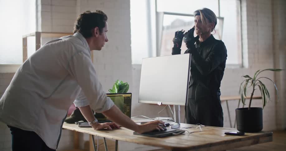 Two men stand in an office while one man wearing virtual reality equipment moves and throws punches as the other man watches his computer as he instructs the man how to move
