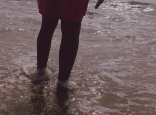 A black man wearing orange shorts is standing on a beach with his feet in shallow water as the waves roll in
