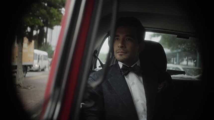 A man wearing a tuxedo suit is sitting in a car and traveling on a city street