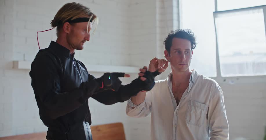 A man has interactive computer devices on his hands and another man shows him hand movements